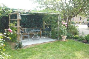 A wooden pergola structure with a table and chairs underneath, surrounded by a garden with various plants and flowers. - Holiday apartment with 2 bedrooms (Sehestedt)