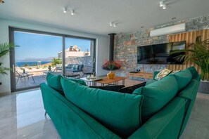 Uma sala de estar moderna com um sofá verde, uma TV de tela plana na parede de pedra e vista para o mar através de grandes janelas.