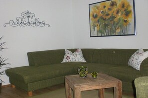 Ein grünes Sofa mit floralen Kissen, ein Couchtisch aus Holz mit Dekorationselementen und ein Sonnenblumenbild an der Wand.