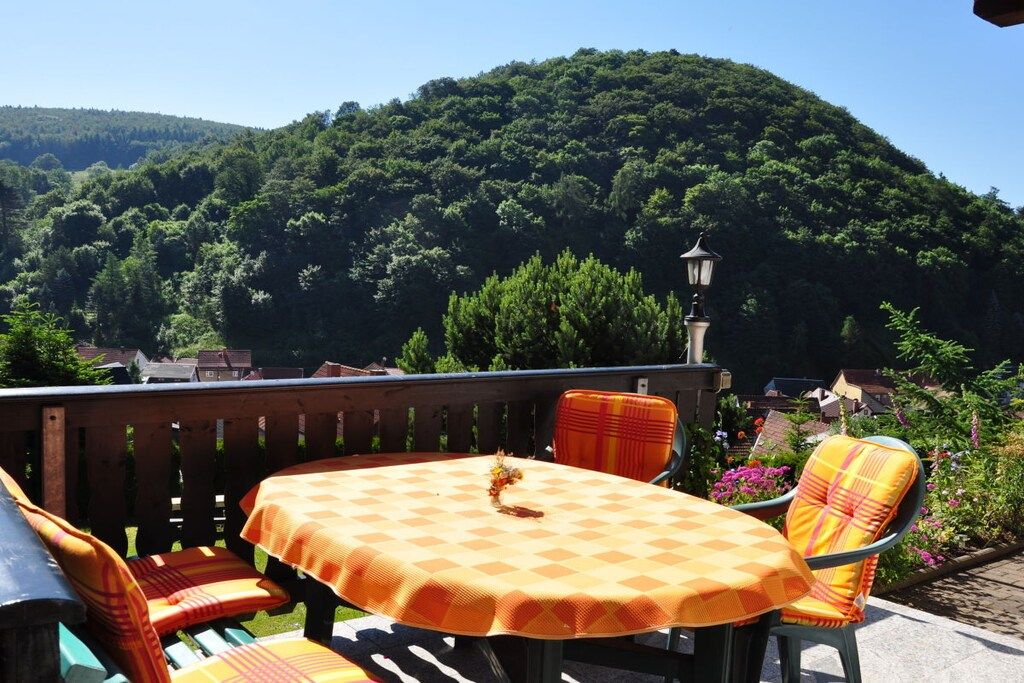 An outdoor patio with a table and chairs, surrounded by a wooden deck and a view of a forested hill.