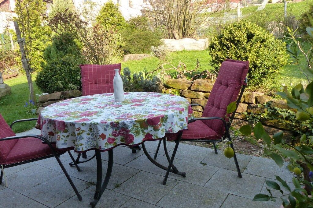 Un patio al aire libre con una mesa redonda y cuatro sillas, rodeado de follaje y un muro de piedra.
