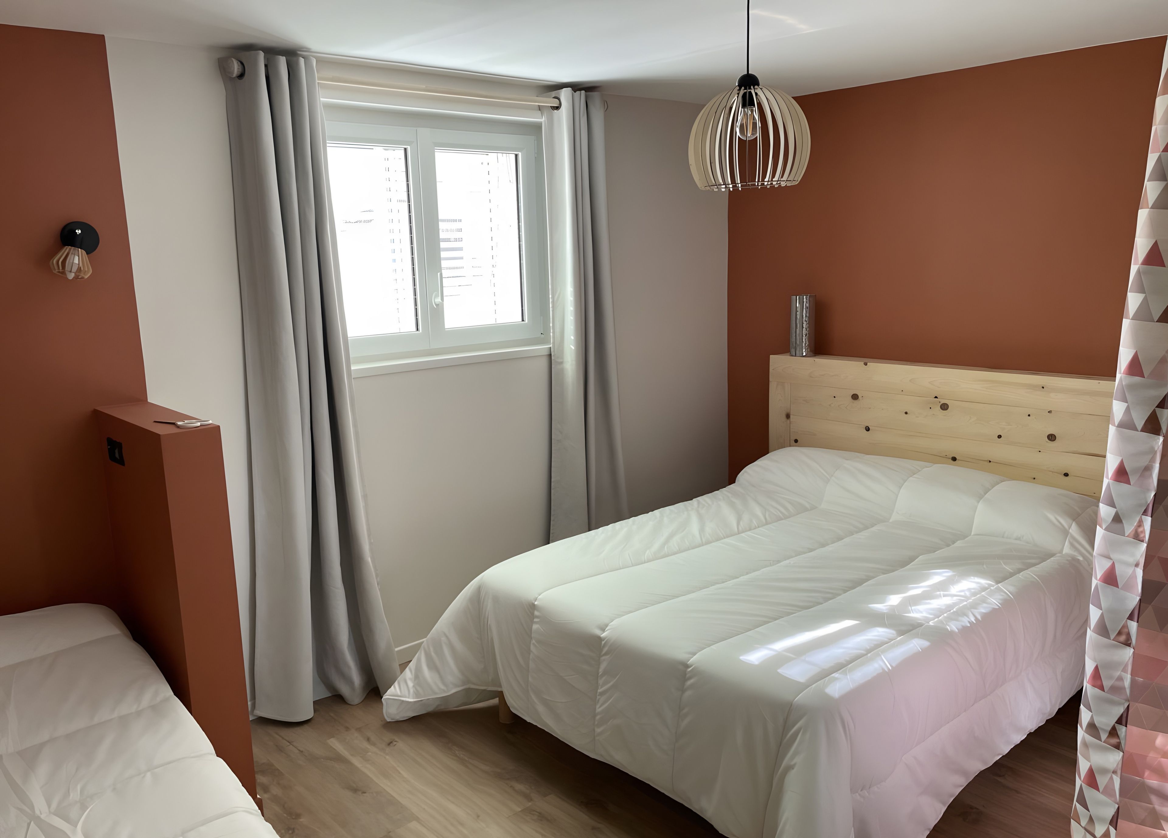 Ein Schlafzimmer mit einem Bett, einem Fenster mit Vorhängen, einer Wandleuchte und einem Holzkopfende.