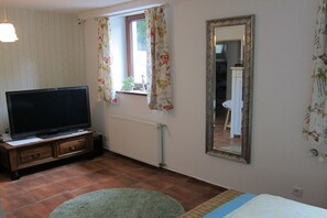 A room with a television, a radiator, a mirror, and a window with curtains. - Heideblockhaus Harmonie (Neuenkirchen)