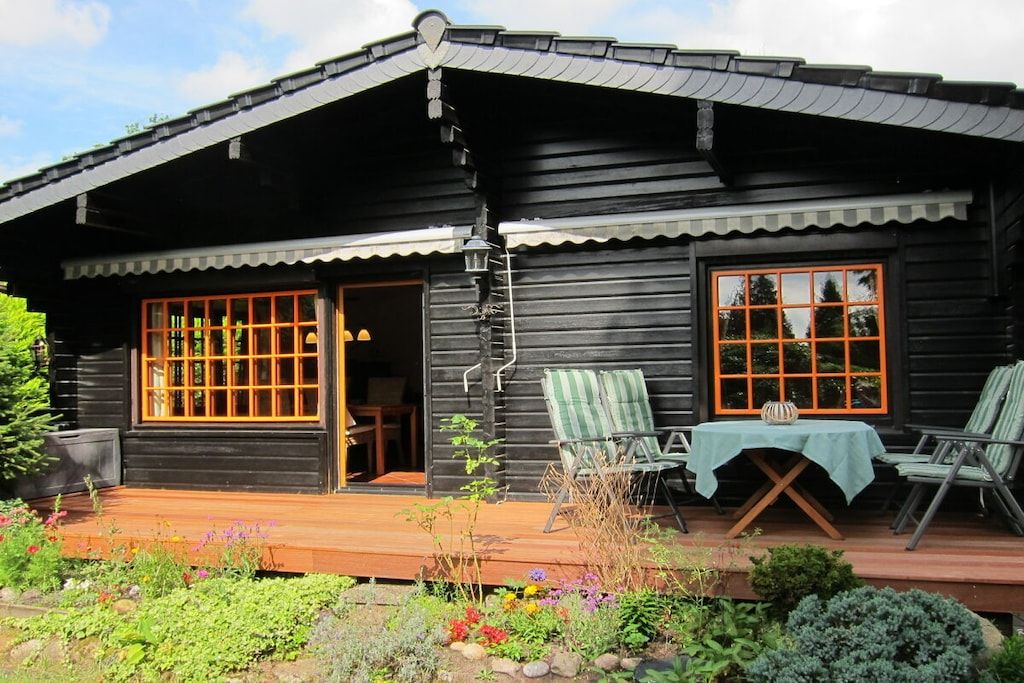 Een houten chalet met een veranda, twee ramen met oranje kaders en een kleine tafel met stoelen.
