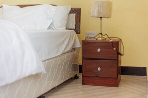 Un lit bien fait, avec du linge de lit blanc, une table de chevet en bois avec une lampe et une prise électrique au mur.