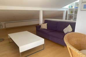 A room with a white table, a purple sofa, a wicker chair, and a bookshelf. - Studio apartment Diessen (Diessen)