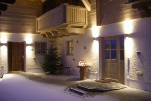 Ein verschneiter Außenbereich mit einem hölzernen Balkon, einer verschneiten Tür und einem kleinen Tisch mit einer Lampe.