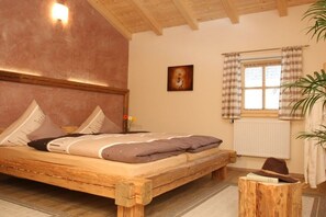 A wooden bed with pillows and a blanket, a small wooden stool with a hat, a radiator, and a window with curtains. - Holiday apartment with 1 bedroom (Freyung)
