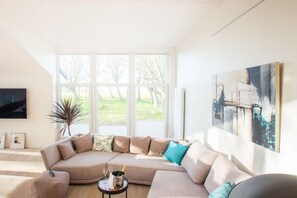 En stue med en hjørnesofa, et fladskærms TV, et stort vindue med udsigt til træer og et billede på væggen.