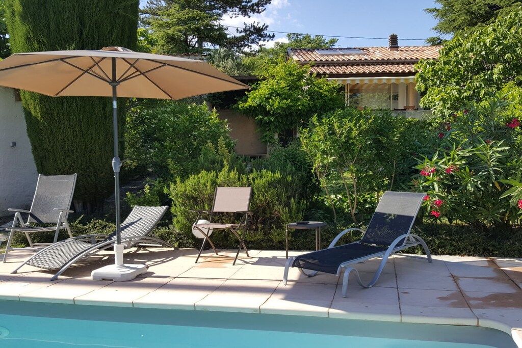 A poolside area with lounge chairs and an umbrella, surrounded by greenery and a building with a tiled roof.