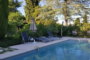 Una piscina con sillas de descanso y sombrillas, rodeada de follaje y árboles.