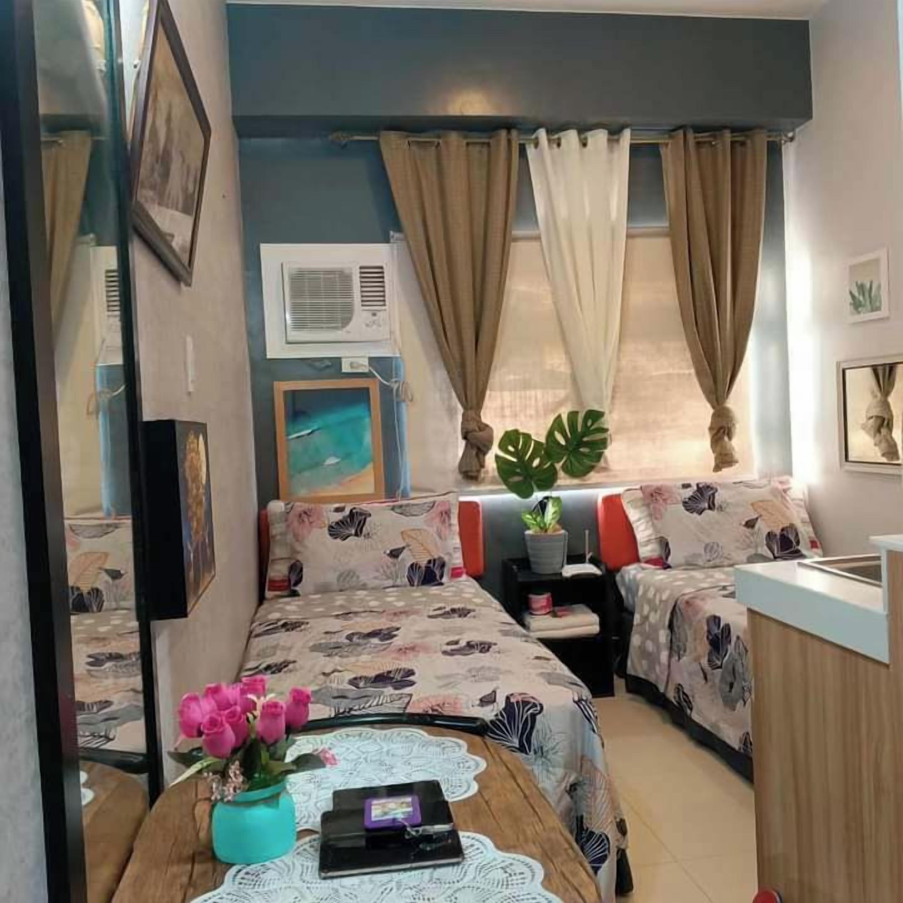 Une pièce de taille modeste comprenant un lit, une petite table ornée d’un vase de fleurs, une fenêtre avec des rideaux et un climatiseur fixé au mur.