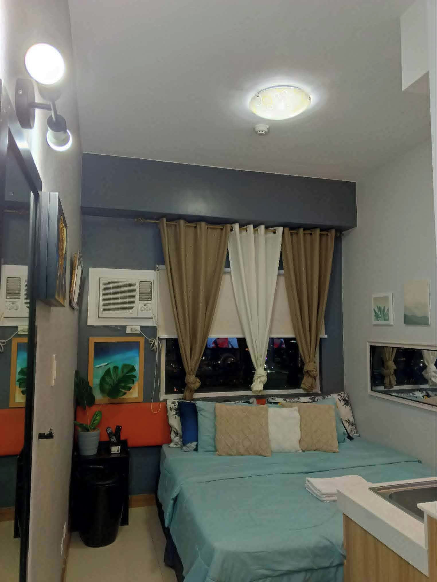A compact bedroom with a bed, a window with curtains, a bedside table, a wall-mounted air conditioner, and a ceiling light.