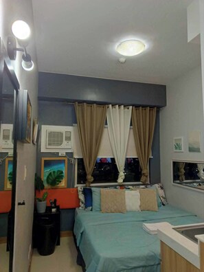 A compact bedroom with a bed, a window with curtains, a bedside table, a wall-mounted air conditioner, and a ceiling light. - Deca Luxe Stay (Mandaluyong)