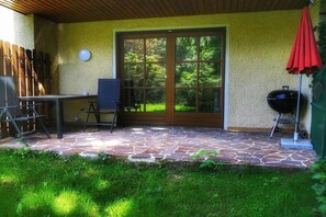 Ein möblierter Terrassenbereich mit Steinboden, einem Holztisch, einem Grill und einer Tür zu einem Garten.
