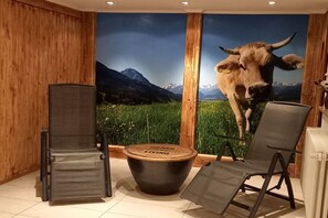 A room with a large wall mural of a cow, a black folding chair, a small round table, and a wooden panel wall. - Eldorado Holiday Apartment Southwind (Bolsterlang)