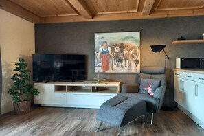 A living room with a flat-screen TV, a painting, a sofa, a coffee table, a lamp, and a kitchen area with cabinets and a microwave. - Eldorado Holiday Apartment Southwind (Bolsterlang)
