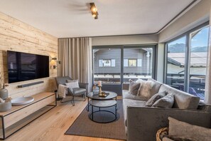 Ein modernes Wohnzimmer mit einem flachbildfernseher an einer Holzwand, einem grauen Sofa, einem grauen Sessel, einem runden Couchtisch und einem großen Fenster mit Blick auf Gebäude und Berge.