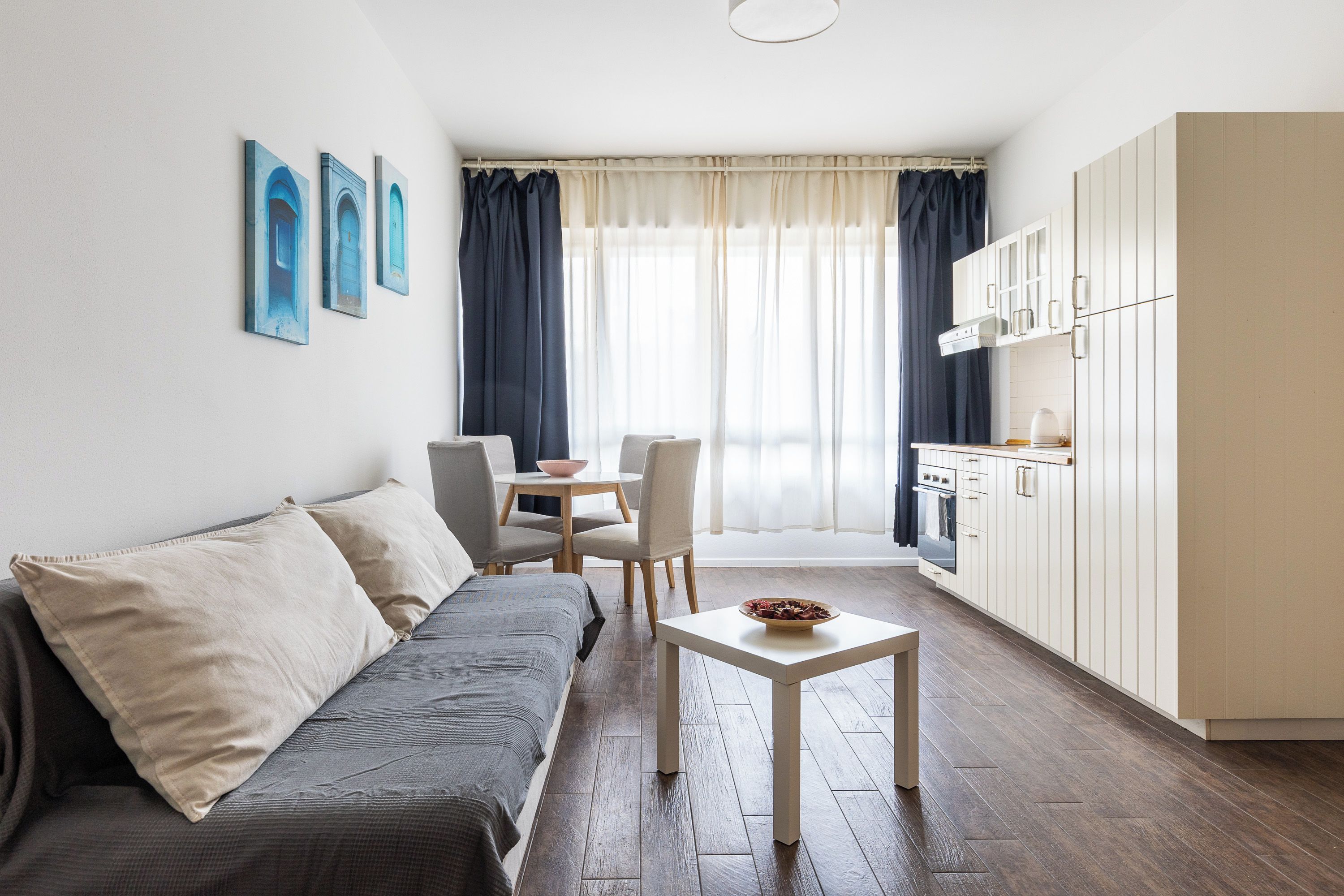 Un espacio habitable compacto con sofá, una mesa de comedor con sillas, una pequeña cocina y un ventanal con cortinas.