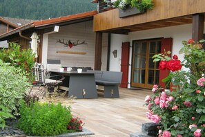 Eine Holzhütte mit Essbereich, einer Couch und einem Tisch mit Stühlen. Ein Balkon mit Pflanzen und einem roten Strauch ist vorhanden.