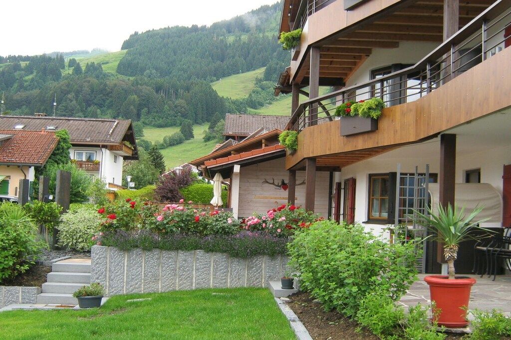 Un edificio de varios pisos con balcones, un jardín con diversas plantas y flores, y una vista de colinas verdes al fondo.