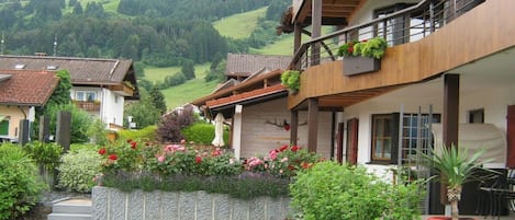 Un edificio de varios pisos con balcones, un jardín con diversas plantas y flores, y una vista de colinas verdes al fondo.