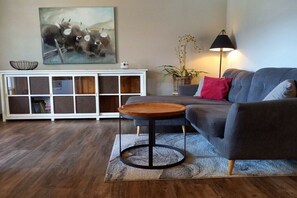 Un salon avec un canapé gris, une table basse ronde en bois, un meuble blanc avec des portes en verre, un lampadaire et un tableau représentant des taureaux accroché au mur.