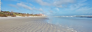A beach with a building in the background, sandy shore, and ocean waves. - Relax in a spacious 1BR wooded retreat in New Smyrna Beach with a modern open kitchen, comfortable living area and covered back deck overlooking nature. (New Smyrna Beach)