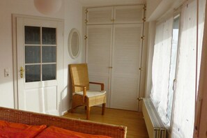 Um quarto com porta fechada, uma cadeira de vime, um radiador e uma janela com cortinas.