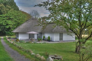 A house with a thatched roof, a paved driveway, and a well-maintained garden with a variety of plants and a seating area. - Holiday home in Sörup with swimming pool (Sörup)