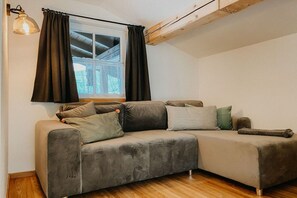 A room with a grey sofa, a window with curtains, a wooden ceiling, and a wall-mounted lamp. - Holiday apartment with 1 bedroom (Schönau am Königssee)