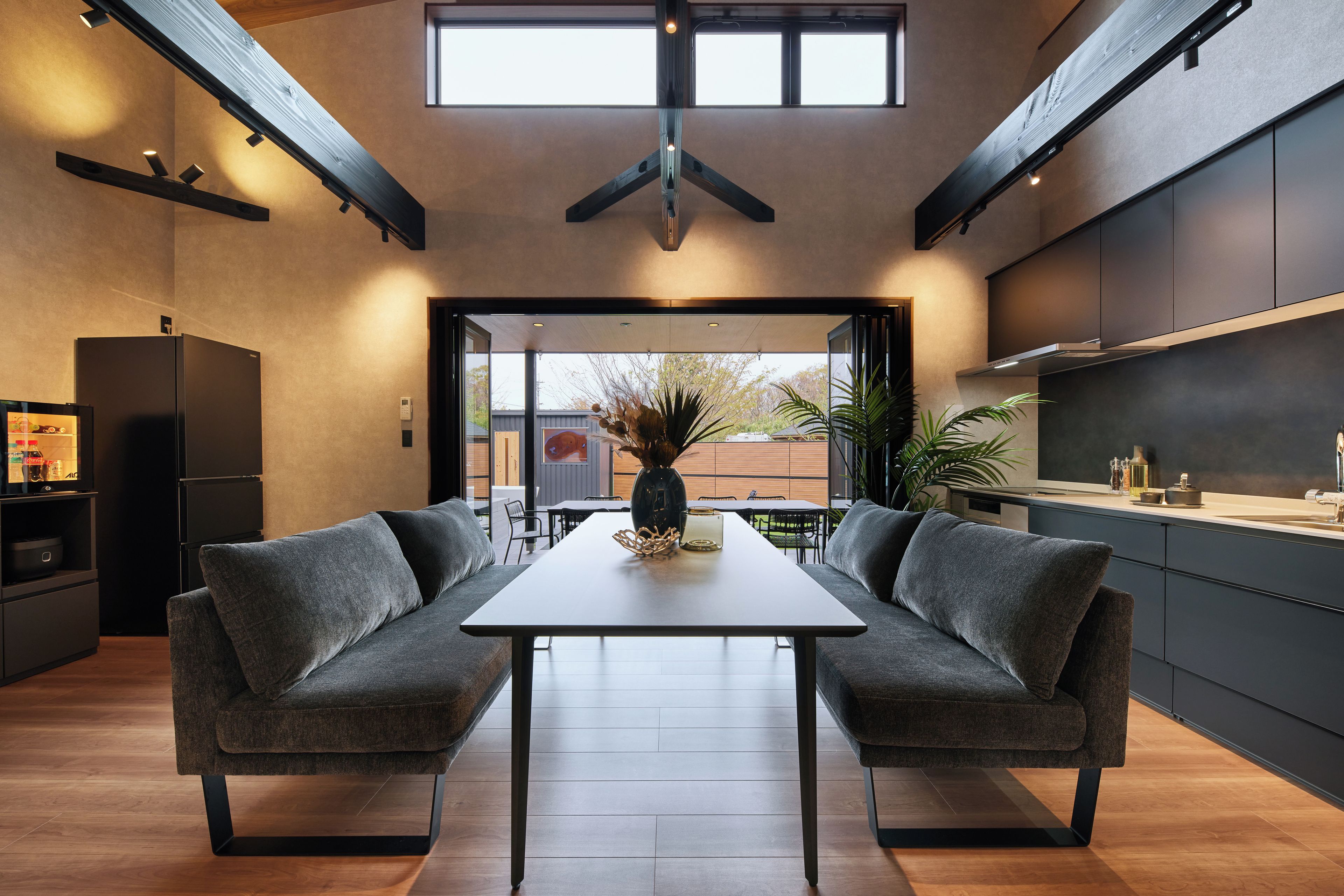 A modern kitchen with a dining area, featuring a central table with chairs, a refrigerator, and a large window providing natural light.