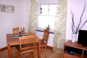 Ein Zimmer mit einem Holzespeisetisch, zwei Stühlen, einem kleinen Topfpflanzen, einem Fernseher auf einem Holzständer und einem Fenster mit Blick nach draußen.