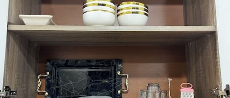 A kitchen cabinet with two shelves, one holding white bowls with gold bands and the other with a black marble backsplash, a white sink, and various kitchen utensils.