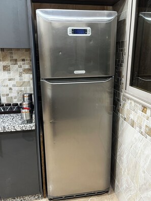 A stainless steel refrigerator with a digital display, located in a kitchen with tiled backsplash and granite countertops.