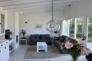 Ein modernes Wohnzimmer mit einem weißen Sofa, einem weißen Couchtisch und einer Chandelüster. Ein Fernseher steht auf einer weißen Kommode, und ein kleiner Tisch mit Lampe und Blumen.