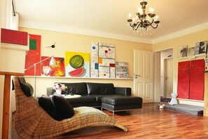 A living room with a black leather sofa, a wicker chair, a chandelier, and abstract wall art. - Holiday Lounge & Art (Bad Driburg)