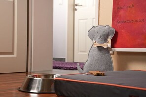 A grey plush toy with a tag on a black cushion with orange trim, a metal dog bowl, and a red wall with text. - Holiday Lounge & Art (Bad Driburg)