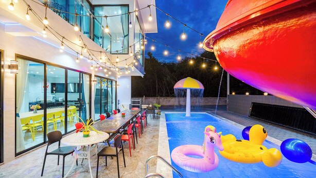 An outdoor pool area with a dining table and chairs, surrounded by a modern building with large windows and string lights. - Love U Pool Villa-Hua Hin (Tambon Hua Hin)