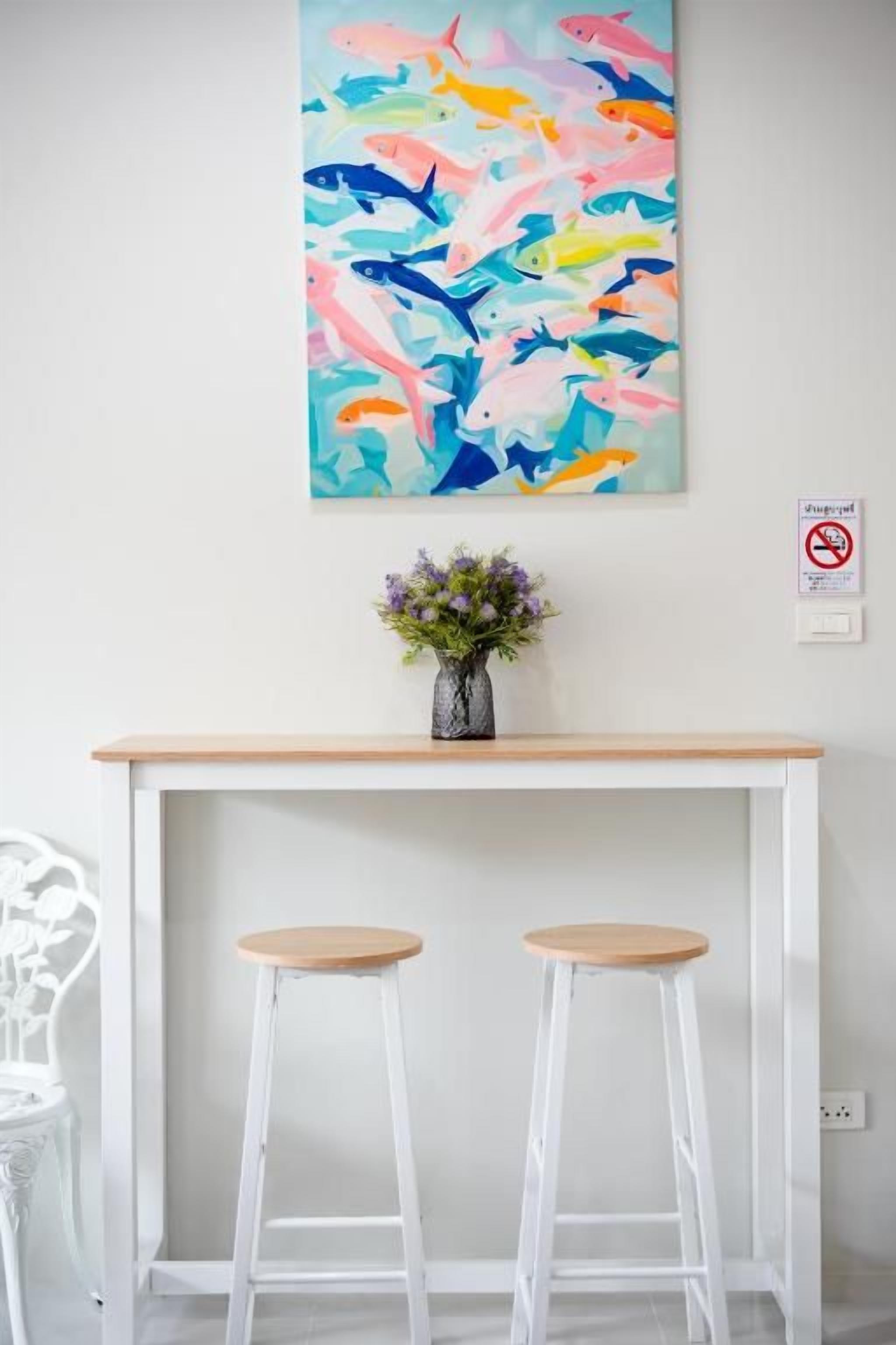 A room with a white desk, two white bar stools, a colorful fish painting, and a small vase with flowers.