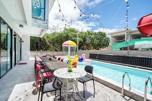 Área de piscina externa com mesa e cadeiras, cercada por uma grade de proteção e luzes penduradas.