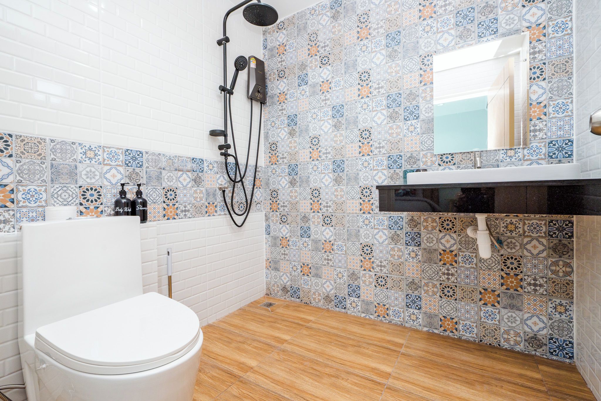 Un baño con inodoro blanco, ducha con cabezal de ducha de mano y lavabo con espejo encima. Las paredes están cubiertas de azulejos con un diseño decorativo.