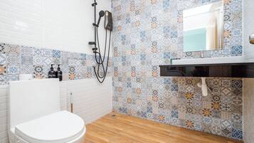 Un baño con inodoro blanco, ducha con cabezal de ducha de mano y lavabo con espejo encima. Las paredes están cubiertas de azulejos con un diseño decorativo.