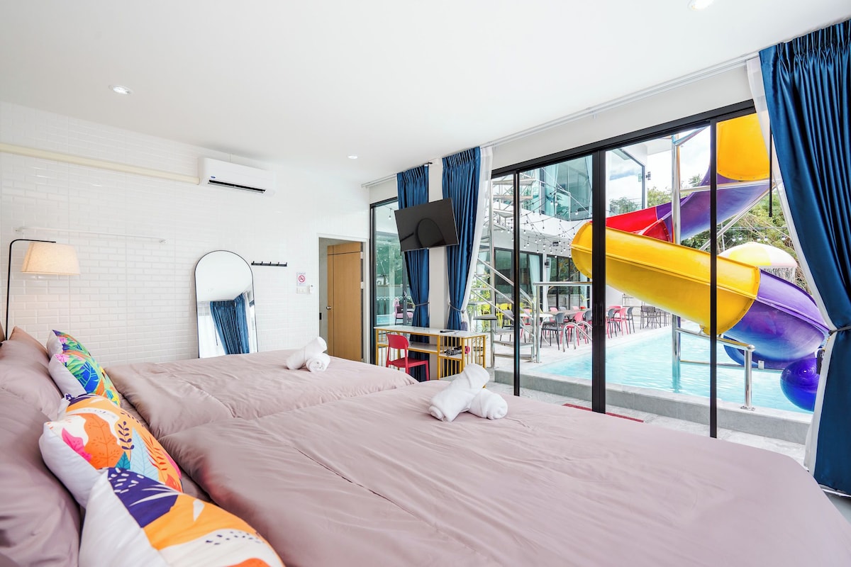 A hotel room with a bed, a desk, a chair, a TV, and a large window with a view of a pool and slides.
