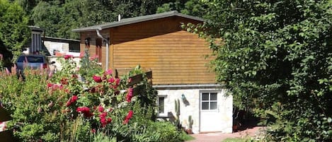 Una cabaña de madera con un sendero de ladrillo que conduce hacia ella, rodeada de vegetación exuberante y plantas en flor.