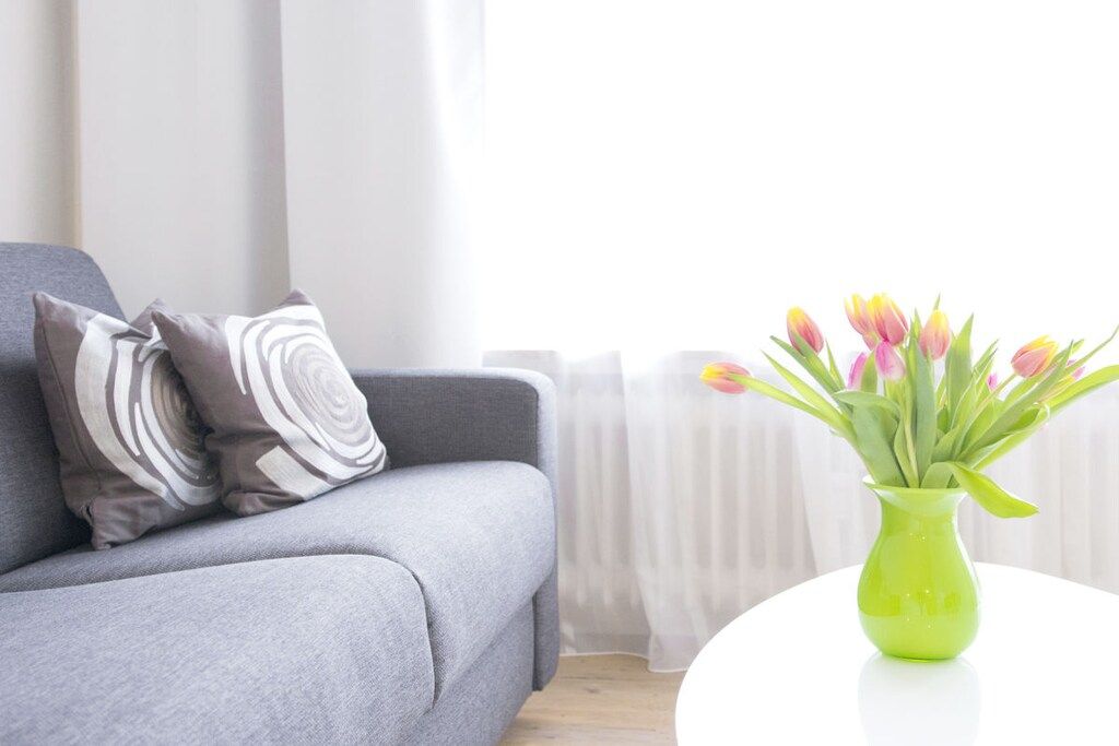 Een grijze bank met gekleurde kussens en een groene vaas met bloemen op een witte tafel.