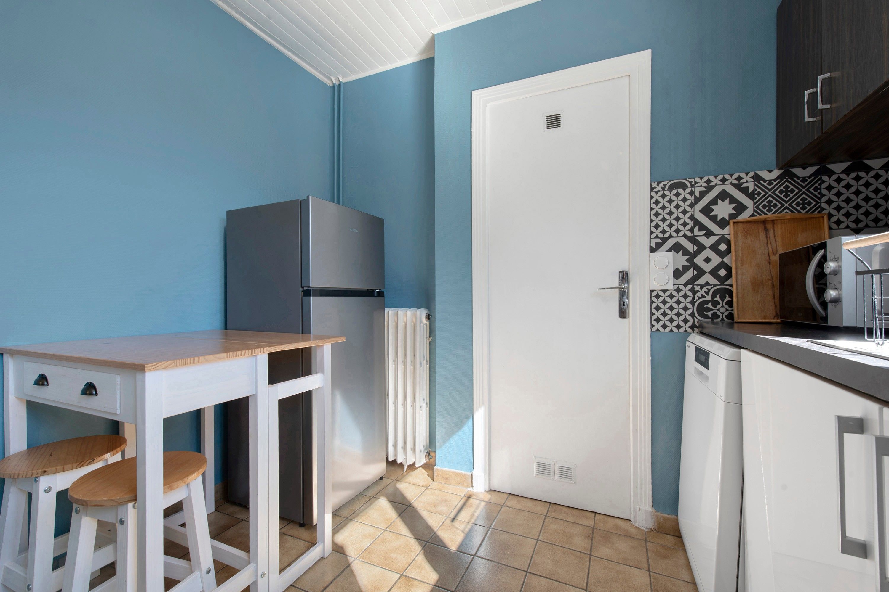 A compact kitchen with a white refrigerator, a wooden table with stools, a white door, and patterned backsplash tiles.