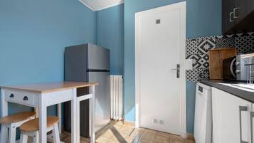 A compact kitchen with a white refrigerator, a wooden table with stools, a white door, and patterned backsplash tiles.