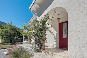 A building with a red door, white walls, and a garden with flowering plants. - Holiday Home 'Chez Négus' with Wi-Fi and Air Conditioning (Laruns)