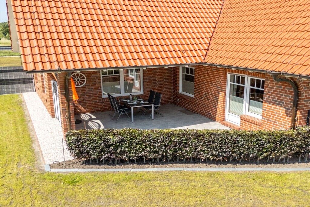 Une maison en briques avec un toit en tuiles, une terrasse avec une table et des chaises, et une haie devant.